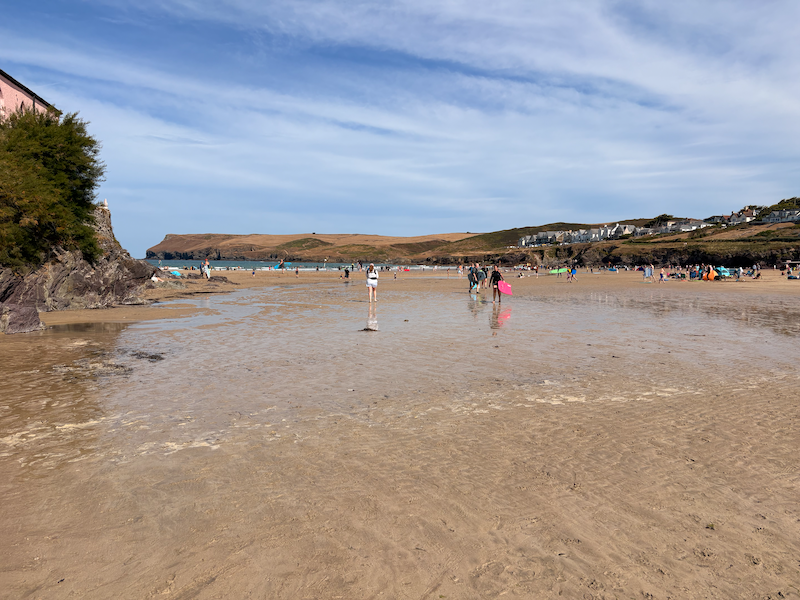 Polzeath Beach, a sandy bay surrounded by cliffs, there are people carrying surfboards and families with windbreaks