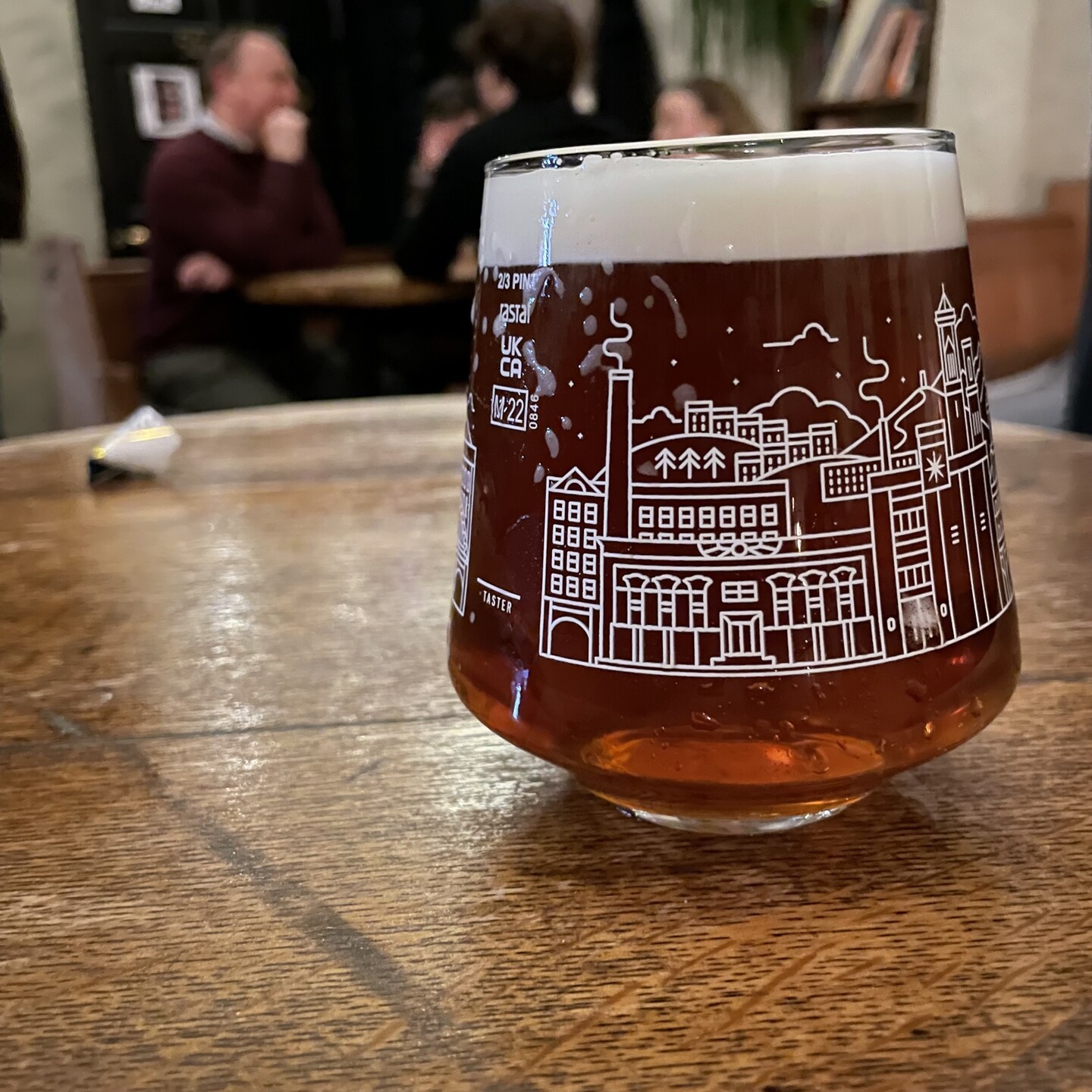 A Northern Monk two-thirds of a pint glass, decorated with white outline drawing which I think might be the brewery building. On an old wooden table in a pub. Containing some Spitting Feathers special ale.
