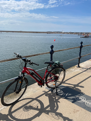 My red eBike parked on the seafront. You can see tiny silhouettes of people on the marine lake and even smaller silhouettes of people walking out to the tidal islands in the estuary.