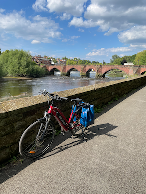 My red eBike with blue shopping bags parked by the river. You can see the River Dee and the stone arches of the Old Dee bridge, built in 1387, in the background; I’d just ridden across it. There’s green trees and a blue sky with not too many white clouds in it.